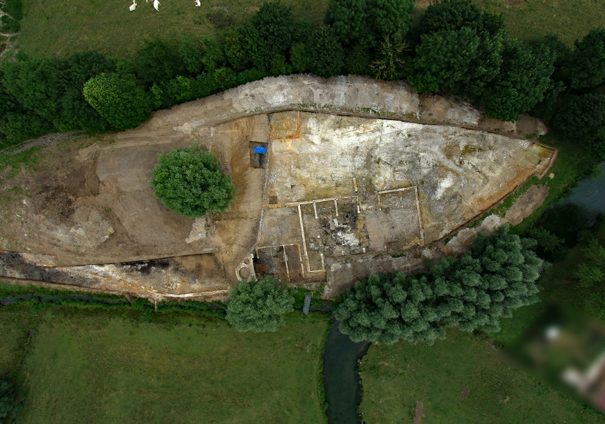 :  Photo de la vue aérienne de l’emprise totale de la fouille de la Cour Lévèque incluant un plan des lieux par transparence. Au centre gauche de la vue aérienne, la cuisine borde la douve. Au-dessus de la cuisine, les vestiges des maçonneries de la résidence regroupant la galerie, la salle d’apparat et les latrines. La résidence est entourée par la douve. Au centre de l’image, un ponton bois permet de traverser la douve pour rejoindre l’espace domestique et agricole. Dans le centre droit de l’image, les écuries et les secondes latrines.