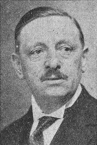 Portrait noir et blanc montrant un homme portant une petite moustache et une cravatte sous un costume sombre.