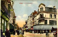 Carte postale en couleurs représentant une rue animée d'Arras où circulent automobiles, cyclistes et passants. On remarque une devanture bleue portant le nom du magasin "À la maison bleue".