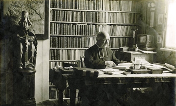 Photographie noir et blanc d'un homme âgé en tenue ecclésiastique assis devant un bureau. Il tient un livre. Derrière lui, une bibliothèque, un crucifix et la statue d'un saint.