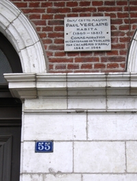 Photographie couleur d'une plaque apposée sur un mur en briques. En-dessous, on remarque un numéro de maison portant les chiffres "55". 