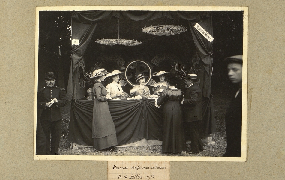 Photographie d’un stand de vente de confiseries tenu par trois femmes dont l’une remplissant des cornets. Décor d’ombrelles chinoises et inscription : "on rend la monnaie". Deux femmes et un homme accoudés au comptoir. Au premier plan, deux hommes dont un soldat dégustant un cornet de friandises. Légende : kermesse des femmes de France. 13-14 juillet 1913.