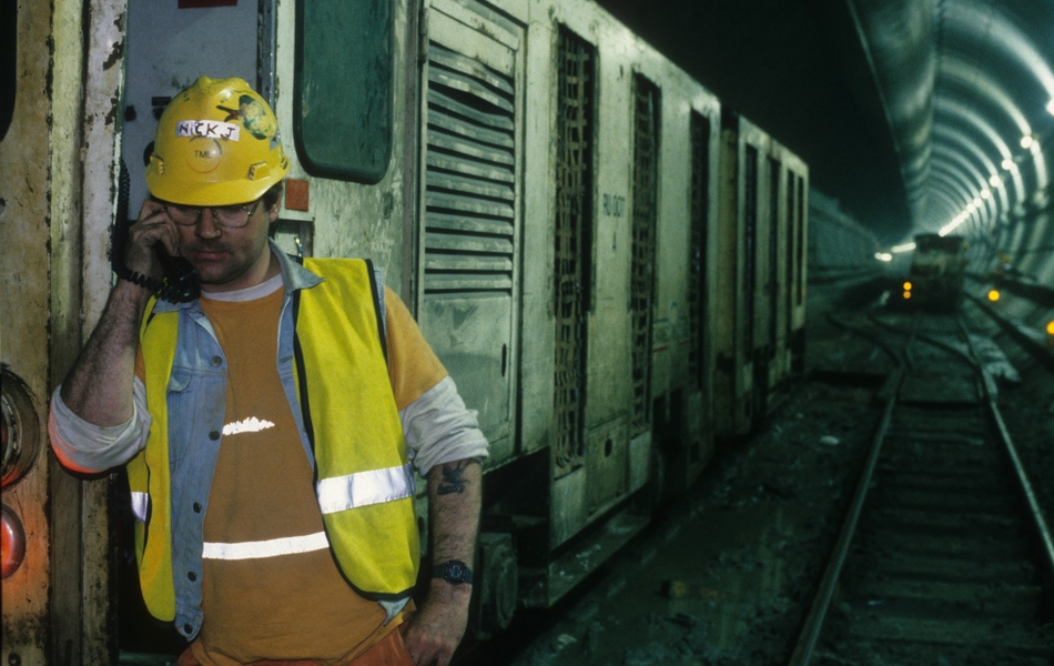 Photographie couleur montrant des rails et des wagonnets dans un tunnel. Au premier plan, un ouvrier adossé à un wagon et portant un casque jaune téléphone.