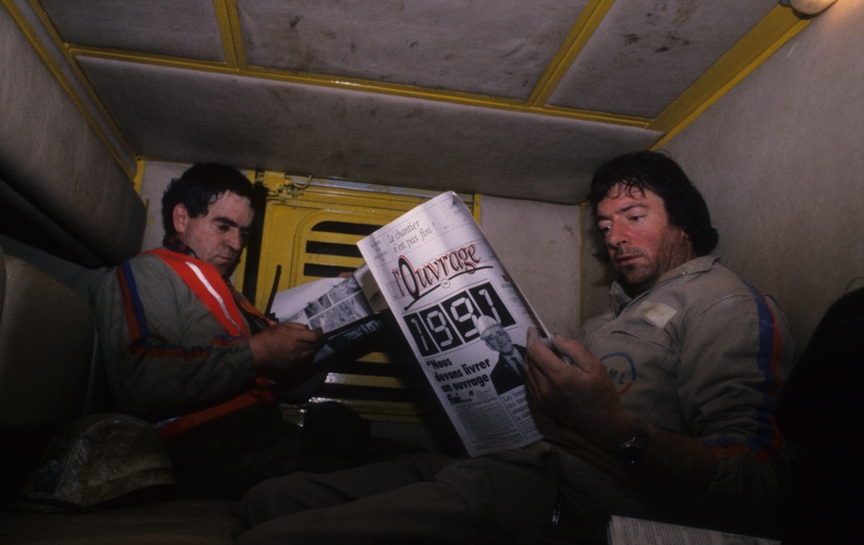Photographie couleur montrant deux ouvriers assis, en train de lire un journal sur lequel on lit "L'ouvrage. Le chantier n'est pas fini!".