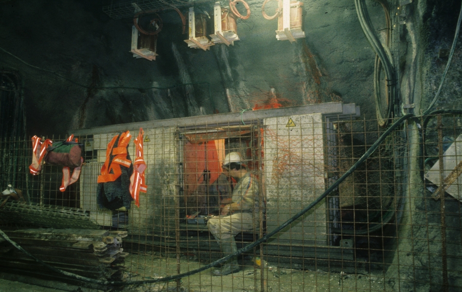 Photographie couleur montrant un wagonnet dans un tunnel, entouré de matériel de chantier. À l'intérieur, deux ouvriers sont assis.