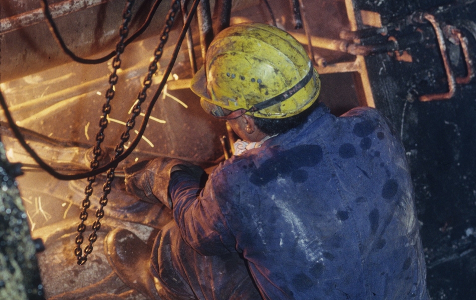 Photographie couleur montrant un ouvrier de dos en train de travailler. Il est vêtu d'une tenue de protection, d'un casque de chantier jaune, de bottes et de lunettes de protection. Des étincelles jaillissent d'une structure métallique sur lequel il est penché. 