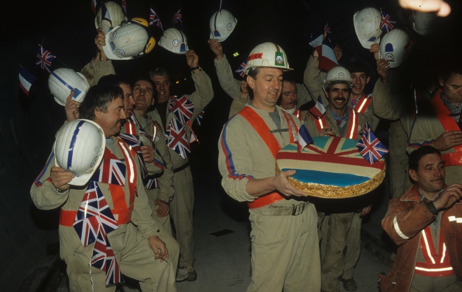 Photographie couleur montrant des ouvriers souriant, levant leur casque et prés de drapeaux britanniques. Au premier plan, un ouvrier porte un gâteau aux couleurs de la France et du Royaume-Uni.