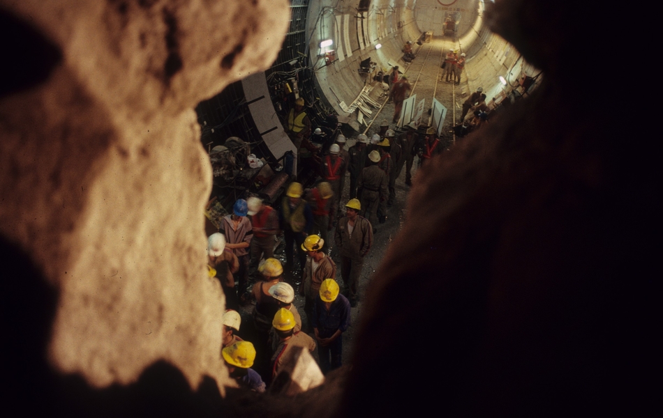 Photographie couleur prise d'un interstice dans une paroi. On voit en contre-bas des ouvriers dans un tunnel.