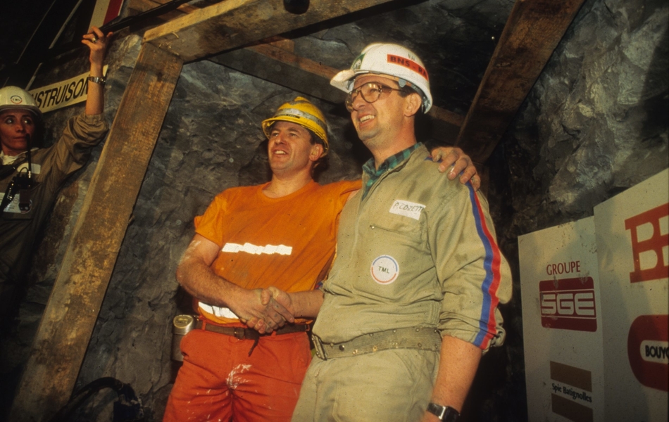 Photographie couleur montrant deux hommes en tenue d'ouvrier se serrant la main dans une galerie de tunnel.
