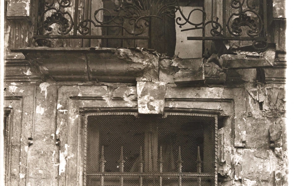 Photographie noir et blanc montrant un balcon ouvragé en fer, fort endommagé.