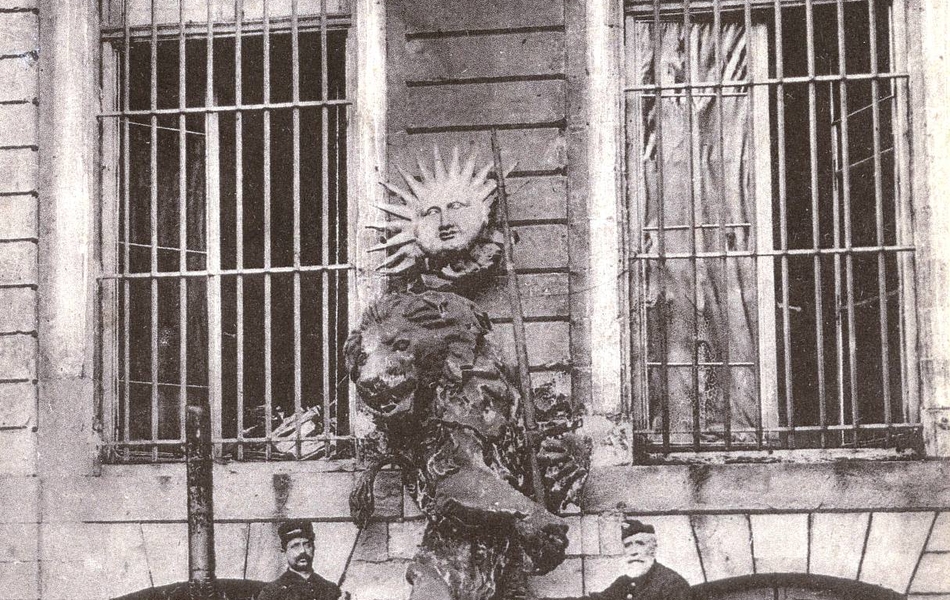 Carte postale noire et blanc montrant les vestiges d'une statue de lion, posée contre un mur, encadré par deux hommes.