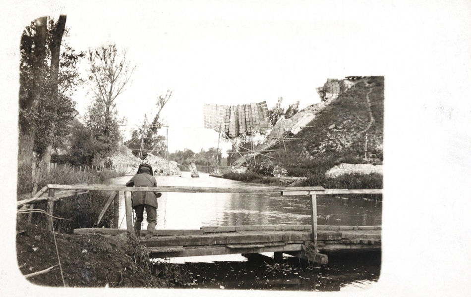 Photographie noir et blanc montrant au premier plan un homme de dos sur un pont au-dessus d'une rivière, photographiant un système de camouflage se trouvant sur l'autre rive.