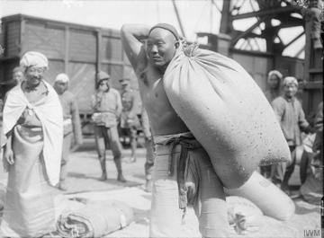 Photographie noir et blanc montrant au premier plan un asiatique, torse nu, portant un sac sur son épaule. À l'arrière plan, d'autres hommes devant un wagon de marchandises.