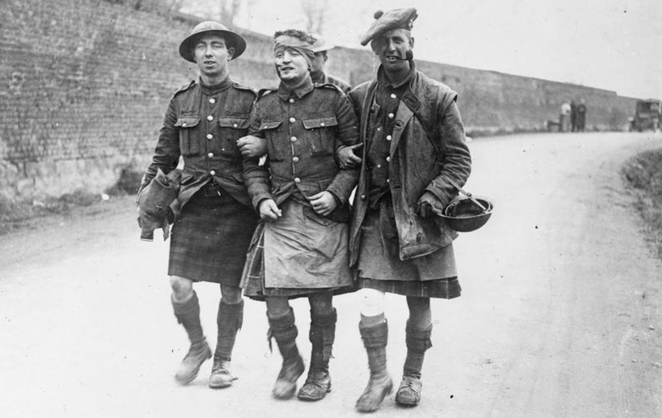 Photographie noir et blanc montrant deux hommes en kilt soutenant un troisième qui semble blessé à la tête.