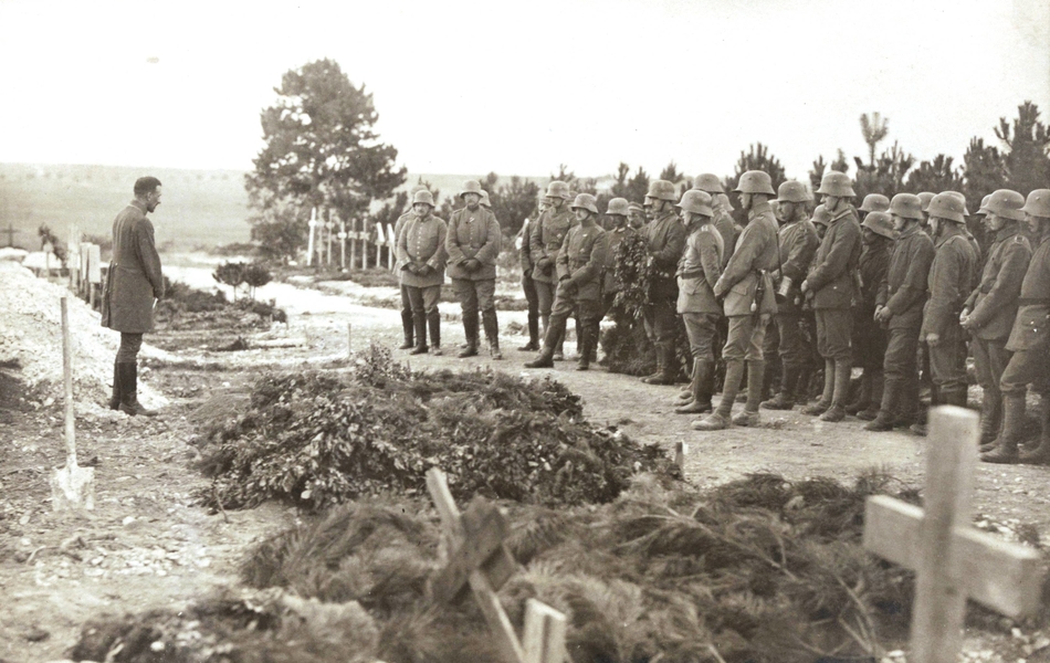 Photographie noir et blanc montrant un homme recueilli devant une tombe fraîchement remblayée. Derrière se trouve un groupe d'hommes dans la même posture.