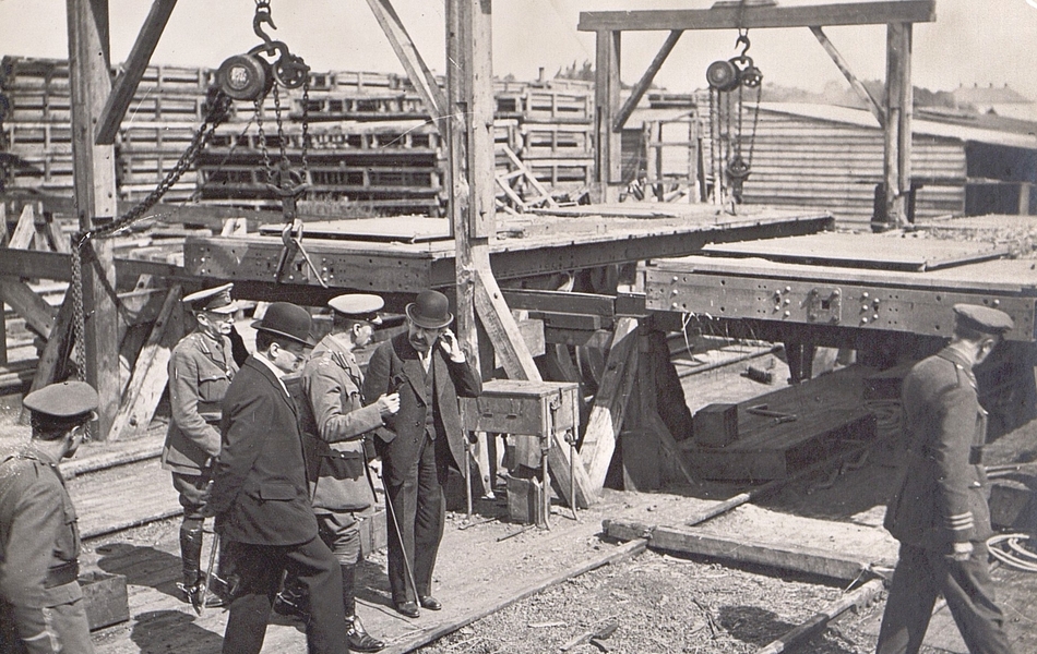 Photographie noir et blanc montrant deux civils entourés de militaires en train de discuter autour de machines et de matériel.