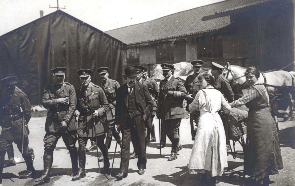 Photographie noir et blanc montrant une délégation de soldats accompagnant un homme habillé d'un costume, qui croisent deux femmes poussant une brouette pleine de paquets.