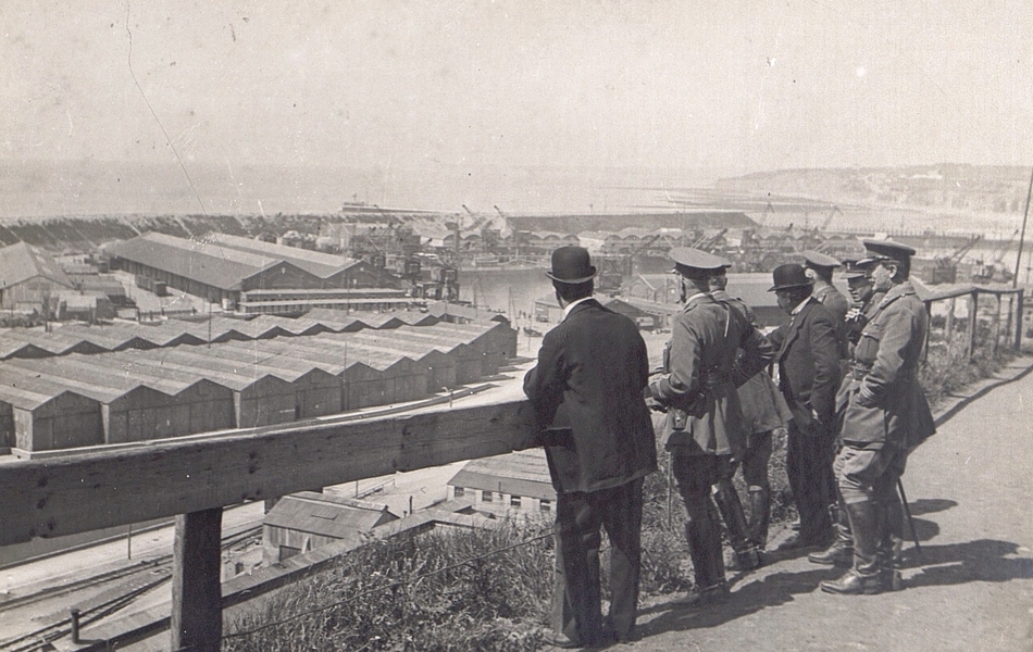 Photographie noir et blanc montrant deux civils entourés de militaires de dos, regardant des entrepôts en contre-bas.