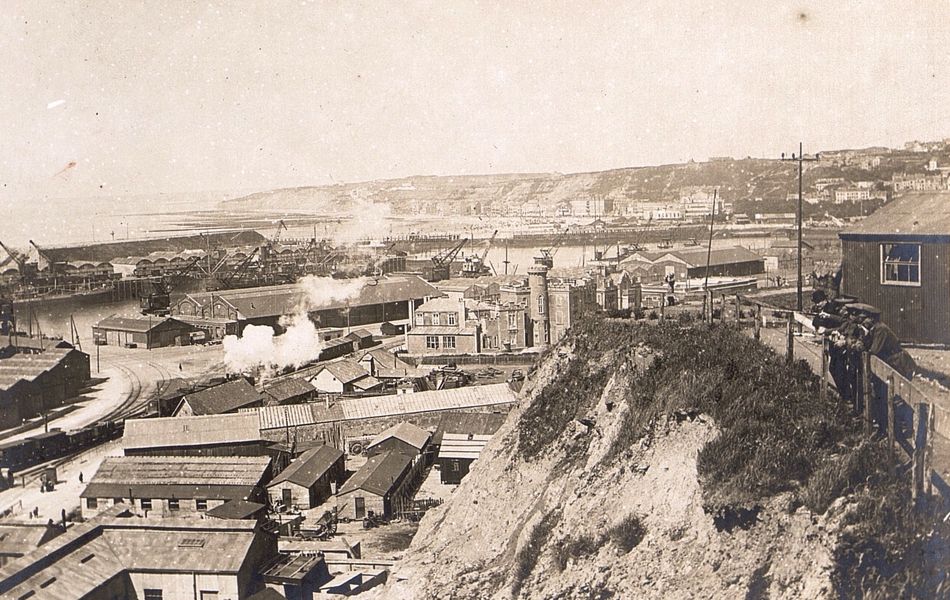 Photographie noir et blanc montrant des entrepôts et des baraquements devant un port.