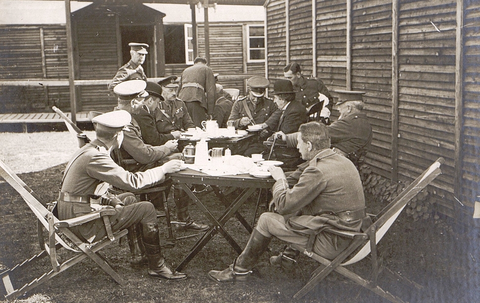 Photographie noir et blanc montrant deux civils et des militaires assis autour de tables devant des baraquements et en train de boire du thé.