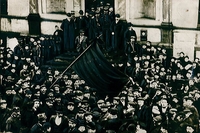 Photographie noir et blanc montrant une foule d'hommes encadrée par la police. Certains brandissent des drapeaux.