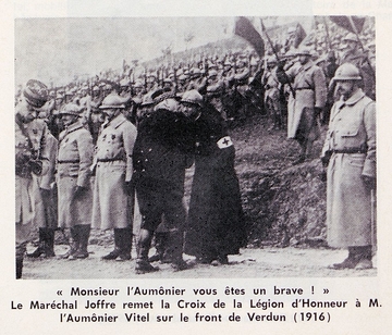 Photographie noir et blanc montrant un soldat embrassant un moine dans une rangée de soldats. En-dessous la légende suivante : "Monsieur l'aumônier, vous êtes un brave ! Le maréchal Joffre remet la croix de la légion d'honneur à M. l'aumônier Vitel sur le front de Verdun, 1916".