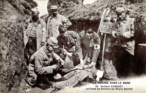 Carte postale noir et blanc montrant un homme se faisant soigner à même le sol dans une tranchée, entouré d'autres. On voit un panneau en bois sur lequel il est écrit "Poste de secours".