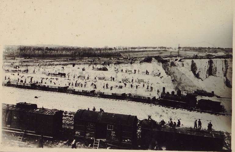 Photographie noire t blanc montrant un chantier dans une plaine. Au premier plan, des rails et des wagons stationnés.