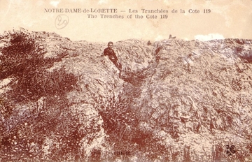 Carte postale sepia montrant les pentes d'une colline striées par des tranchées ; un homme se tient assis dans l'une d'elles.