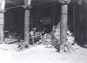 Photographie noir et blanc montrant un soldat assis sous des arcades, en train de lire un journal devant des amas de gravier.