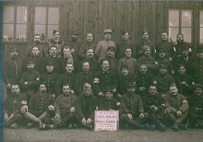 Photographie noir et blanc montrant un groupe d'hommes assemblés autour d'un écriteau (détaillé en légende).
