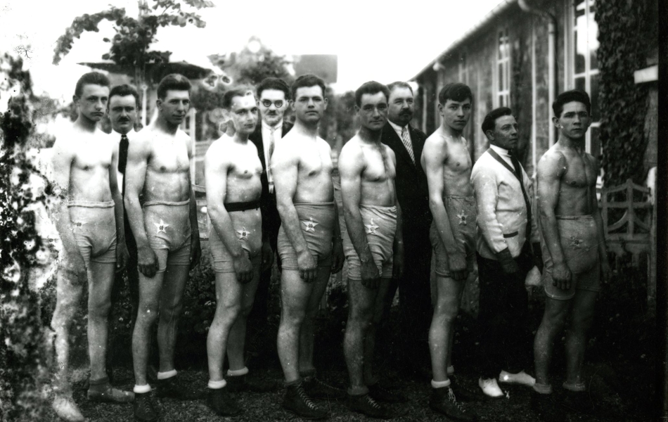 Photographie noir et blanc montrant sept hommes torse nu et en short au premier plan, tournés de profil. Derrière eux se trouvent trois hommes vêtus de costumes.