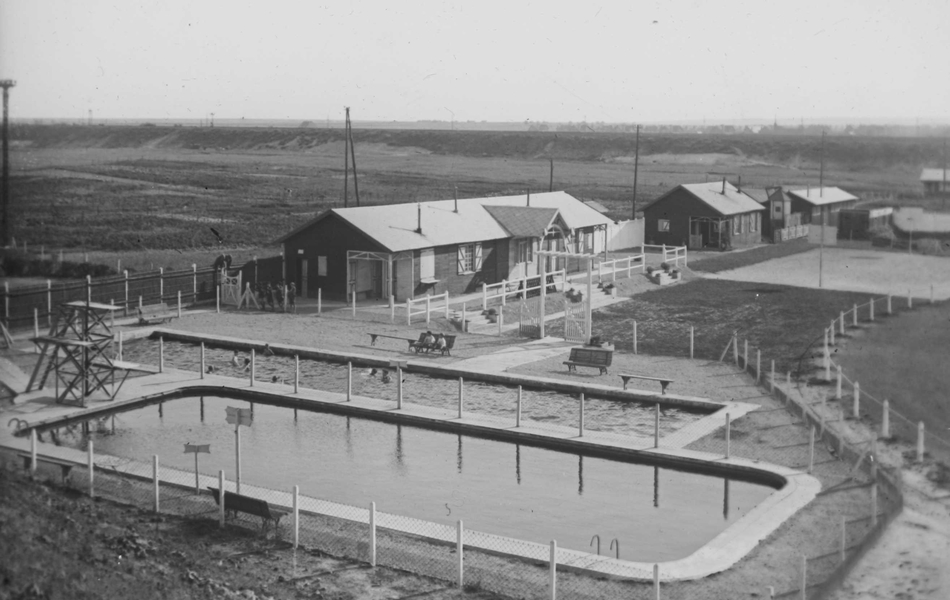 Photographie noir et blanc montrant une piscine extérieure et des bâtiments autour.