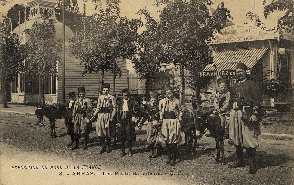 Carte postale noir et blanc de jeunes garçons habillés à la mode mauresque menant des ânes. Ils posent devant des stands, accompagnés d'un homme portant une tenue de zouave.