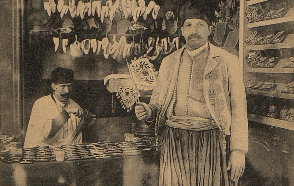 Carte postale noir et blanc montrant l'échoppe d'un artisan oriental. Au premier plan pose un homme en habit traditionnel tenant un éventail.