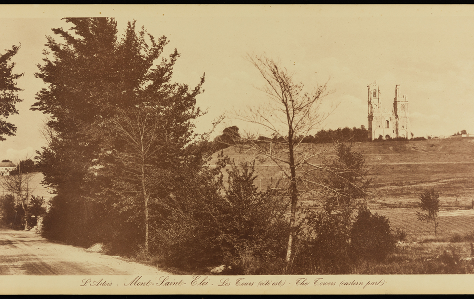 Photographie sépia montrant une route bordant une colline sur laquelle se dresse les ruines de deux tours.