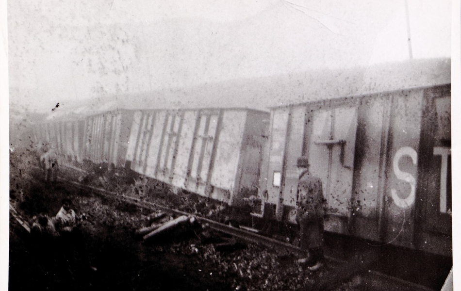 Photographie noir et blanc d'un train déraillé devant lequel passe un homme.