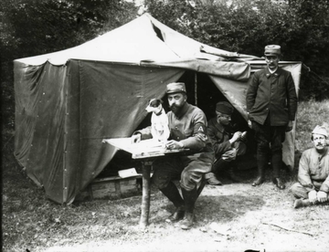 Photographie noir et blanc d'un soldat assis devant une table sur laquelle est posé un petit chien. Derrière, trois autres soldats posent devant une tente.