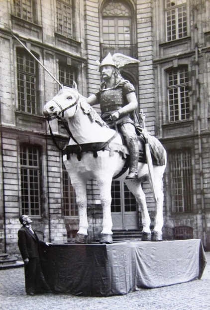 Carte postale noir et blanc du géant Come sur son cheval blanc. Il porte un casque ailé sur de longs cheveux, une grande moustache, une cuirasse et tient dressée une lance. Une épée repose le long de sa jambe gauche.