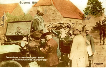 Carte postale colorisée montrant Guillaume II la main devant la bouche, portant le casque à pointe prussien, debout dans une voiture décapotée. À l'arrière de la voiture se trouve un homme assis, en train de boire dans une flasque. Au premier plan, devant la voiture, un groupe d'hommes, dont certains en uniforme militaire allemand.
