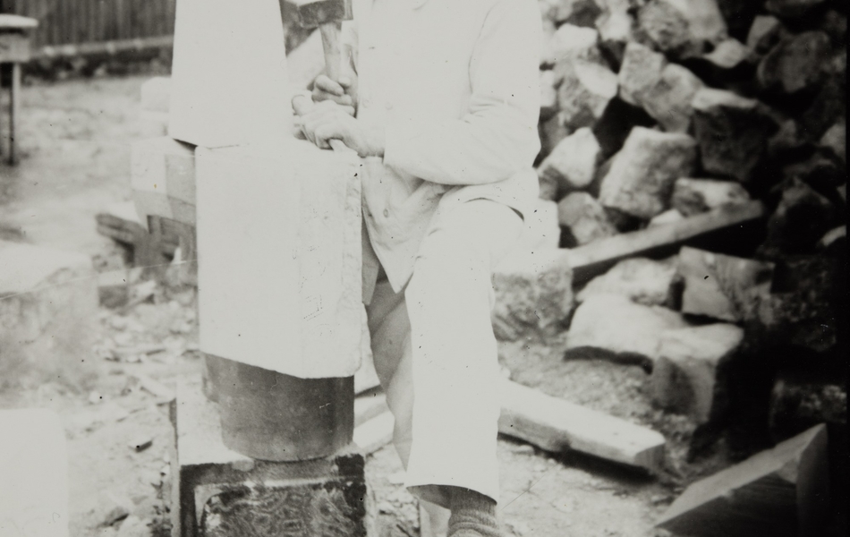 Photographie noir et blanc montrant un homme posant à côté d'une sculpture. À l'arrière on remarque des gravats.