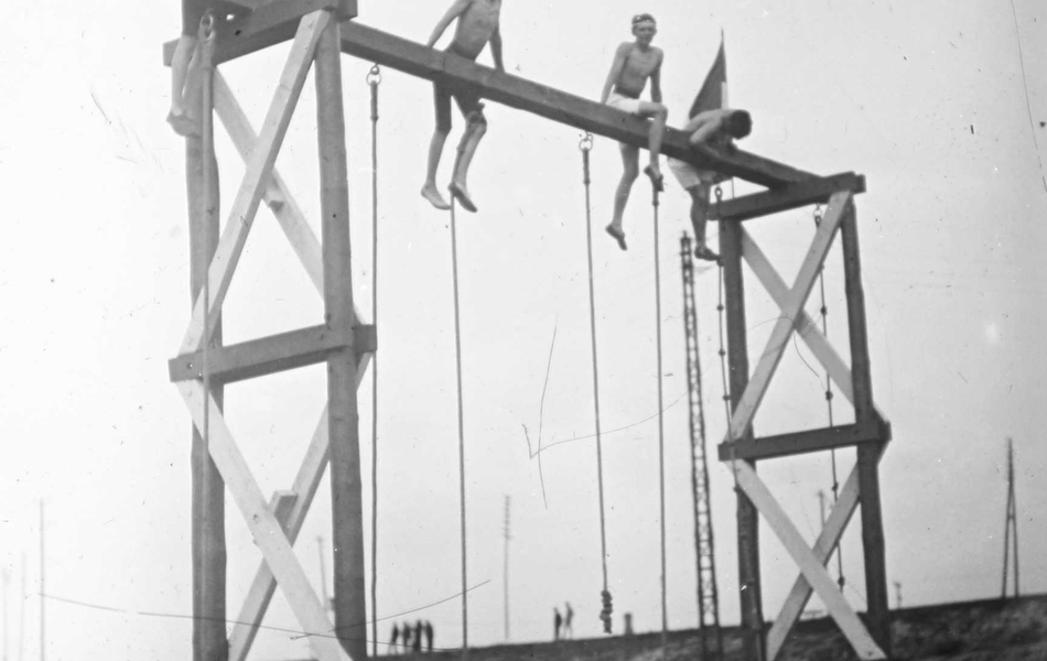 Photographie noir et blanc montrant de jeunes garçons hissés sur uns structure en bois.