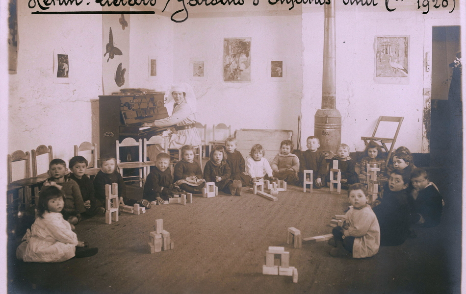 Photographie noir et blanc montrant un groupe de bambins assis par terre en demi-cercle, occupé à jouer avec des cubes en bois.