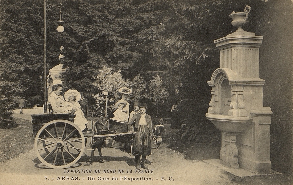 Carte postale noir et blanc d'une carriole attelée à un âne et menée par un garçonnet. L'équipage s'est arrêté devant une fontaine, au détour d'une allée et pose pour le photographe.