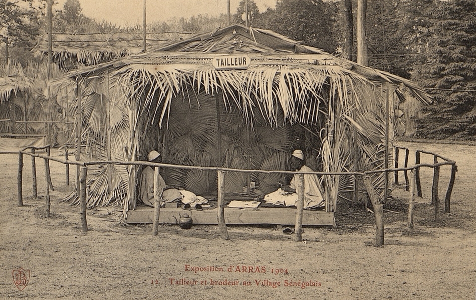 Carte postale noir et blanc de deux hommes assis dans une case ouverte. Au-dessus est accroché le panneau "tailleur".