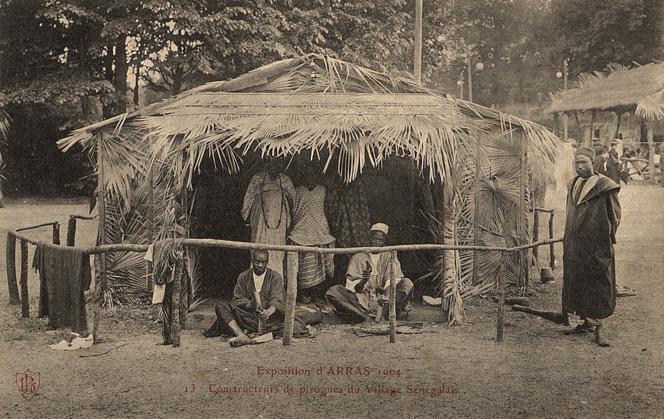 Carte postale noir et blanc d'un groupe d'hommes assis dans une case ouverte et tenant des outils. 