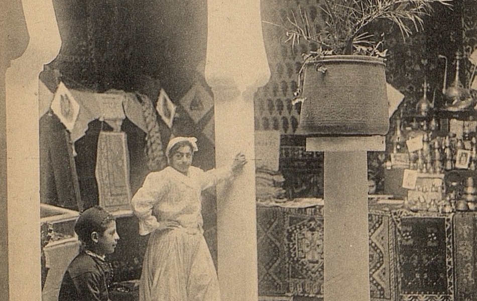 Carte postale noir et blanc montrant une échoppe de tapis et de vaisselle sous une arcade mauresque sur laquelle est appuyée une jeune fille. À ses côtés se trouve un garçon assis sur une chaise.