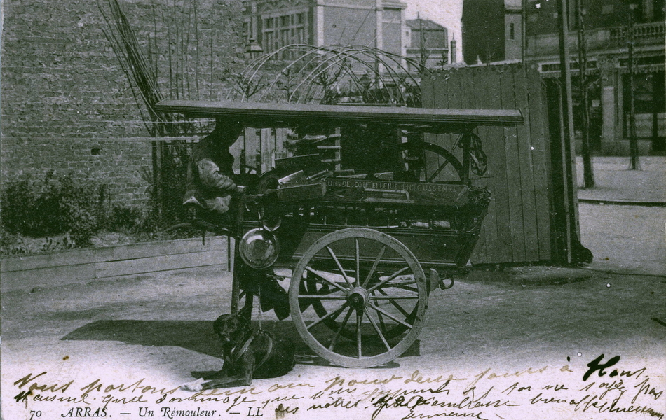 Carte postale noir et blanc d'un homme assis dans une petite carriole chargée de marchandises.