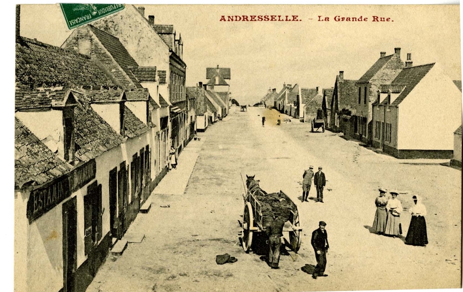 Carte postale en noir et blanc d’une large rue non pavée, bordée de petites maisons mitoyennes. Plusieurs personnes à côté d’une carriole regardent vers le photographe ; au fond de la rue, on devine la mer