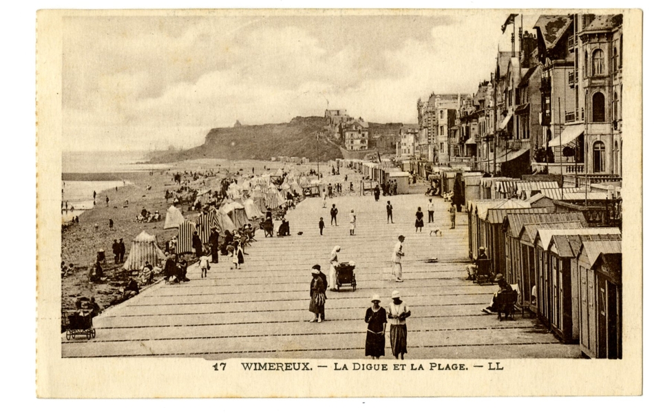 Carte postale en noir et blanc de la digue et de la plage en pleine saison. Des tentes sont installées dans le sable et des cabines de bain le long des maisons sur la promenade